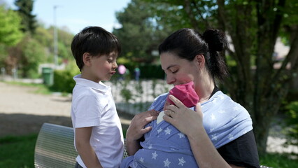 Obraz premium Mother sitting on a bench holding her newborn in a blue star-patterned baby wrap, with a young boy standing beside her, capturing a loving family moment in a suburban park