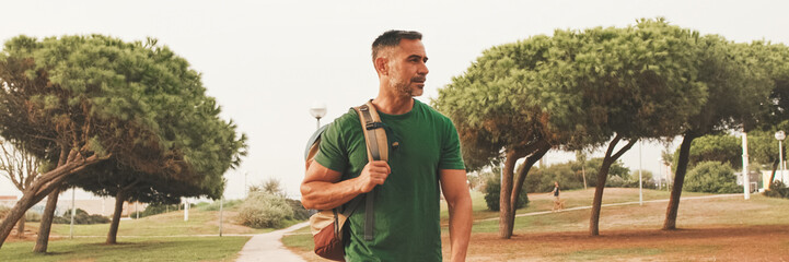 Smiling middle-aged man dressed in casual clothes, with backpack on his shoulder, walks along the...