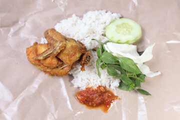 Pecel ayam or ayam penyet is a traditional Javanese food from Indonesia served with fried chicken, rice, cucumber, cabbage, basil and chili sauce isolated on white background, on paper packaging