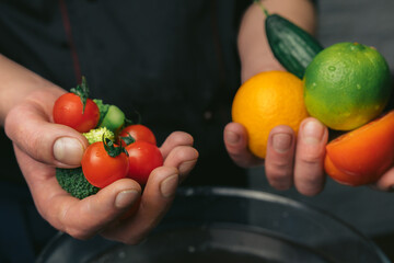 The chef's hands in black jacket hold different vegetables over a bowl of water against the background of a brick wall, ready to wash tomato, cucumber, lemon, lime, broccoli. Space for text.