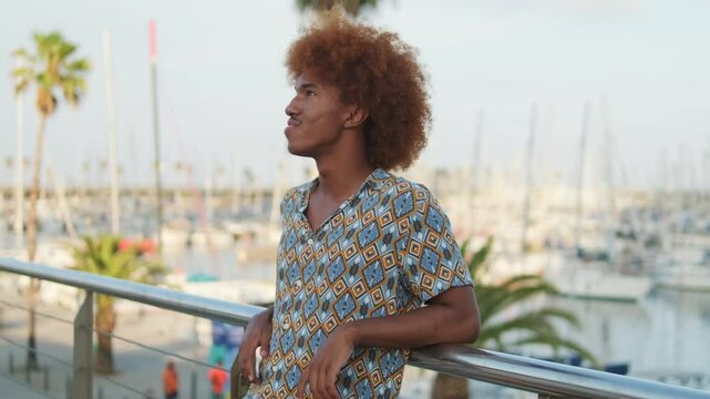 Happy curly-haired guy relaxing against the backdrop oof a yachts and palm trees 