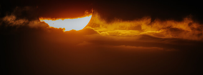 A dramatic sunset with the sun partially obscured by clouds, creating a striking and intense orange...