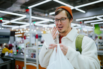 Man wearing beanie and coat looks surprised while examining his receipt at grocery store checkout. Male is shocked by high prices and costs in grocery supermarket, high inflation.