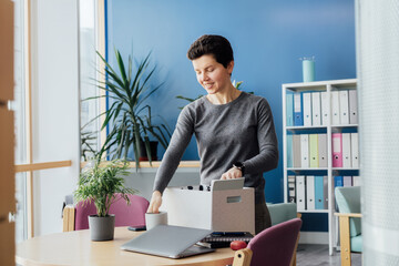 Satisfied newcomer middle aged neutral gender female company worker standing in office unpacking box with personal belongings at new workplace having first working day concept. New Carrier start.