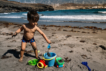 A young boy is playing in the sand on a beach. He is wearing a bathing suit and is holding a bucket. The beach is filled with various toys, including a car and a boat. The scene is lively and playful