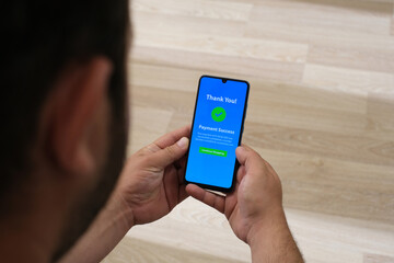 The male hand holding a cell phone with the payment app interface showing the payment successful message with a green check mark. Online shopping, mobile banking and transactions.