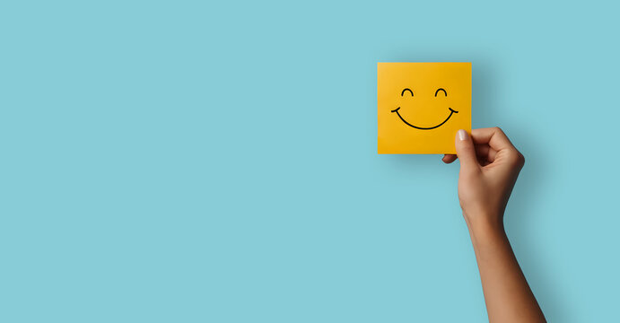 A hand holding a yellow happy smile face on blue background. Positive customer rating, good feedback and review. Happiness and world mental health day concept
