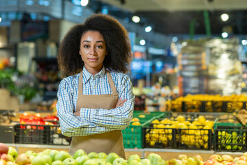Confident young woman working at grocery store, standing with arms crossed among fruits and vegetables. She smiling and wearing an apron. Background showcases colorful produce and market environment