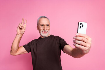 Portrait of cheerful man with beard dressed t-shirt in glasses show v-sign make selfie on smartphone isolated on pink color background