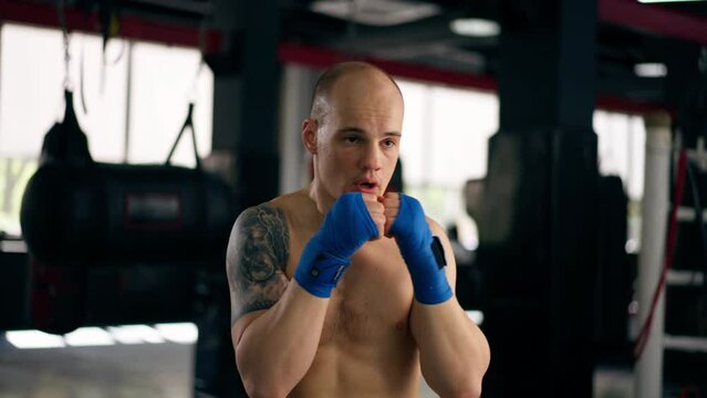 close-up in a boxing fight club bald young boxer with tattoos in blue bandages trains punches shadow boxing professional technique training