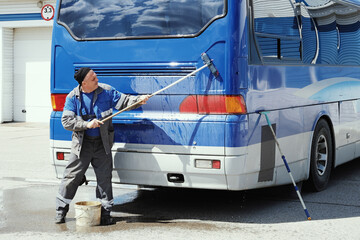 A worker cleans a blue bus exterior with a long-handled brush and water