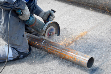 A worker wearing knee pads is cutting a large metal pipe with an angle grinder. The grinder is...
