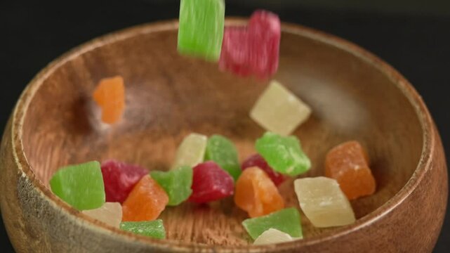 Dried colorful sweet candied fruits mix falling into a wooden bowl in slow motion on black background. Sweets made from dried fruits, kiwi, mango, pineapple, strawberry or gummy candy