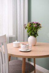 Summer cozy break. Cup of tea, ceramic vaze with flowers on round wooden table near the window.