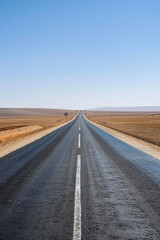 Fototapeta premium Long straight road stretching into the horizon through a vast desert landscape under a clear blue sky.