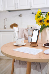 Home workplace in interior - mockup blank white screen of mobile, keyboard, computer mouse, cup of coffee, vase with flowers on wooden table in kitchen.
