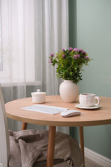  Home workplace in interior - keyboard, computer mouse, cup of coffee, vaze with flowers on wooden table near the window in living room.