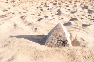 A small stone pyramid partially buried in sandy beach under the sunlight, casting a shadow on the sand.