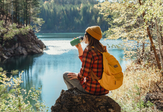 young woman tourist with yellow backpack in hat sitting on stone on shore of lake drinking from thermal mug resting in hike Autumn landscape - Powered by Adobe