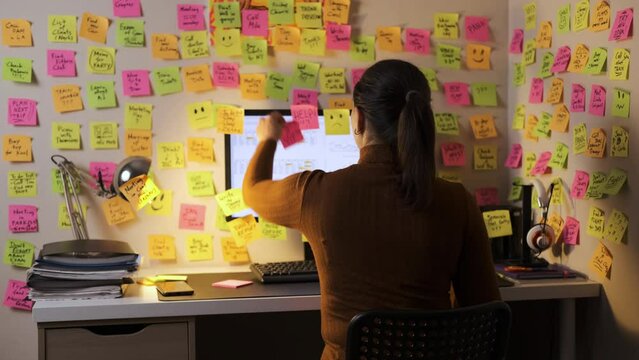 Rear view of young female working late at her desk, adding a colorful paper to her screen, walls filled with lots of vibrant sticky notes with reminders and tasks