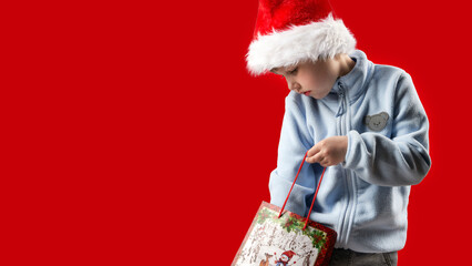 A child wearing a Santa Claus hat pulls a Christmas gift or purchase from a paper bag. The background is red. Copy space.