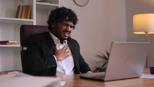 Worried young indian businessman working on laptop while having panic heart attack hold hand on chest and try to calm down at office workplace alone Anxious male with mental health problem indoors