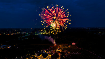 Vibrant Red And Green Fireworks Burst Against The Night Sky, Illuminating A Rural Fairground With White Barns And Small Churches, Enhancing The Festive Atmosphere Amidst The Dark Landscape.