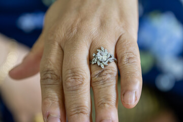 A close-up of a hand adorned with a sparkling diamond ring against a blue background. The elegant...