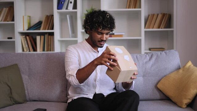 Camera view of positive young indian man influencer looking at camera and talking about shopping in online internet store recording unboxing unpacking delivery box vlog sitting on sofa at home