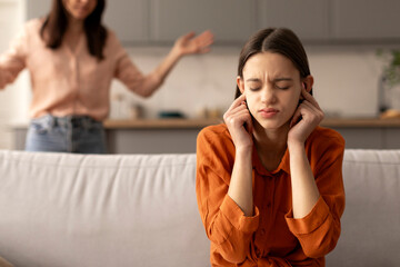 Naughty teen girl covering ears and closing eyes sitting on couch refusing to fulfill mother...