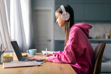 Distance education. Lady teenager in wireless headphones studying online on laptop computer at home, side view. Schoolgirl make homework assignment