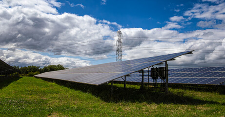 Solar panels in a field with electricity pylon and power lines under a cloudy sky