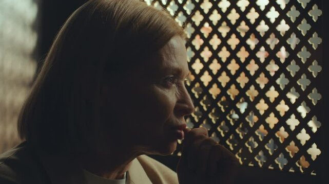 Mature female parishioner having private talk with priest sitting behind latticed opening in confessional booth