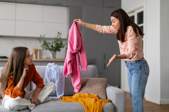 Angry European mother arguing her teen daughter for clothes on sofa, woman holding hoodie and shouting at indifferent girl