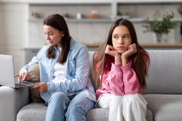 No time for child. Mother working on laptop, sitting on sofa with offended teen daughter nearby,...