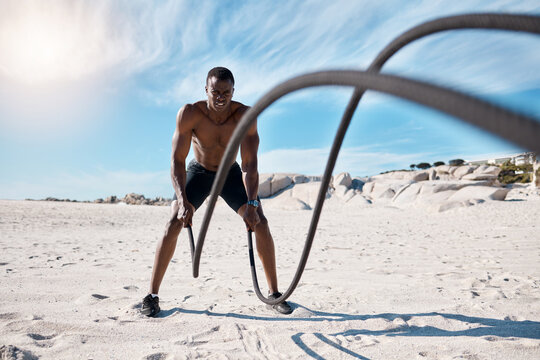 Athlete, fitness and battle ropes on beach for exercise, workout and cardiovascular health outdoor. Black man, wellness and high intensity training by ocean with sun, spiral and slam for strength