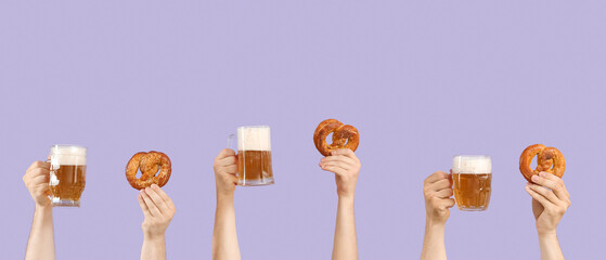 Hands with mugs of beer and pretzels on lilac background