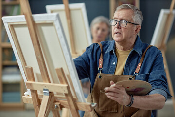 Waist up portrait of creative senior man painting picture on easel in art studio pursuing creative hobby in retirement