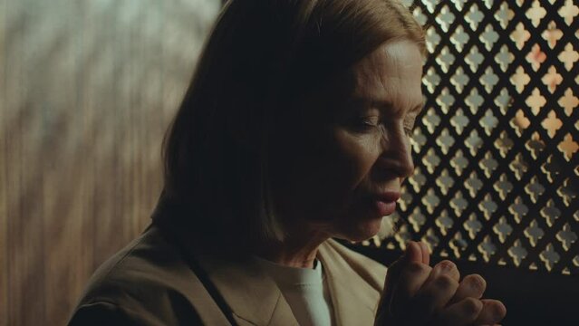 Faithful female parishioner holding palms together and praying to God to absolve sins during confession with priest sitting behind latticed screen
