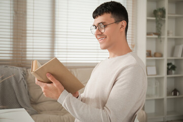Handsome happy young man in new stylish eyeglasses reading book at home