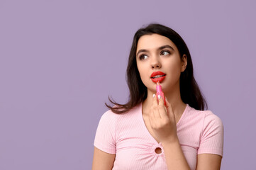 Fototapeta premium Beautiful young woman applying lipstick on lilac background