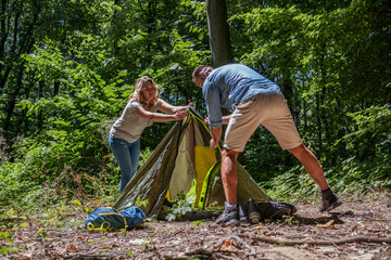 A couple sets up a tent together in nature on a sunny summer day. They work as a team, helping each other to prepare their campsite.