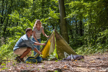 A couple sets up a tent together in nature on a sunny summer day. They work as a team, helping each other to prepare their campsite.