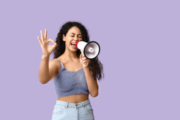 Young African-American woman with megaphone showing OK on lilac background