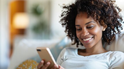 Fototapeta premium A woman with curly hair smiles as she reads something on her phone while sitting comfortably on a couch; the background is soft and warmly lit, creating a cozy atmosphere.