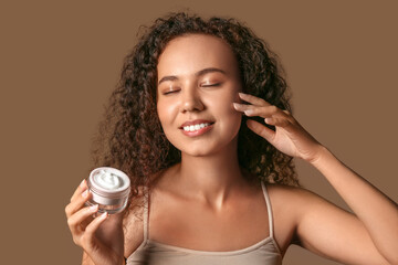 Beautiful young African-American woman with jar of face cream on brown background