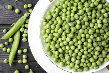 Fresh green peas in bowl and pods on black wooden table, flat lay