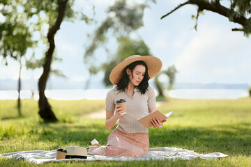 Pretty young woman with cup of coffee reading book on blanket in park