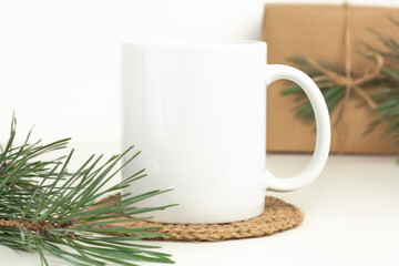 Christmas mockup of white coffee mug on table with jute coaster, pine branches and gift box. Blank 11 oz cup mock up for new year and winter holidays.