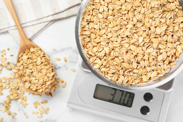 Kitchen scale with bowl of dry oats and spoon on white marble table, flat lay